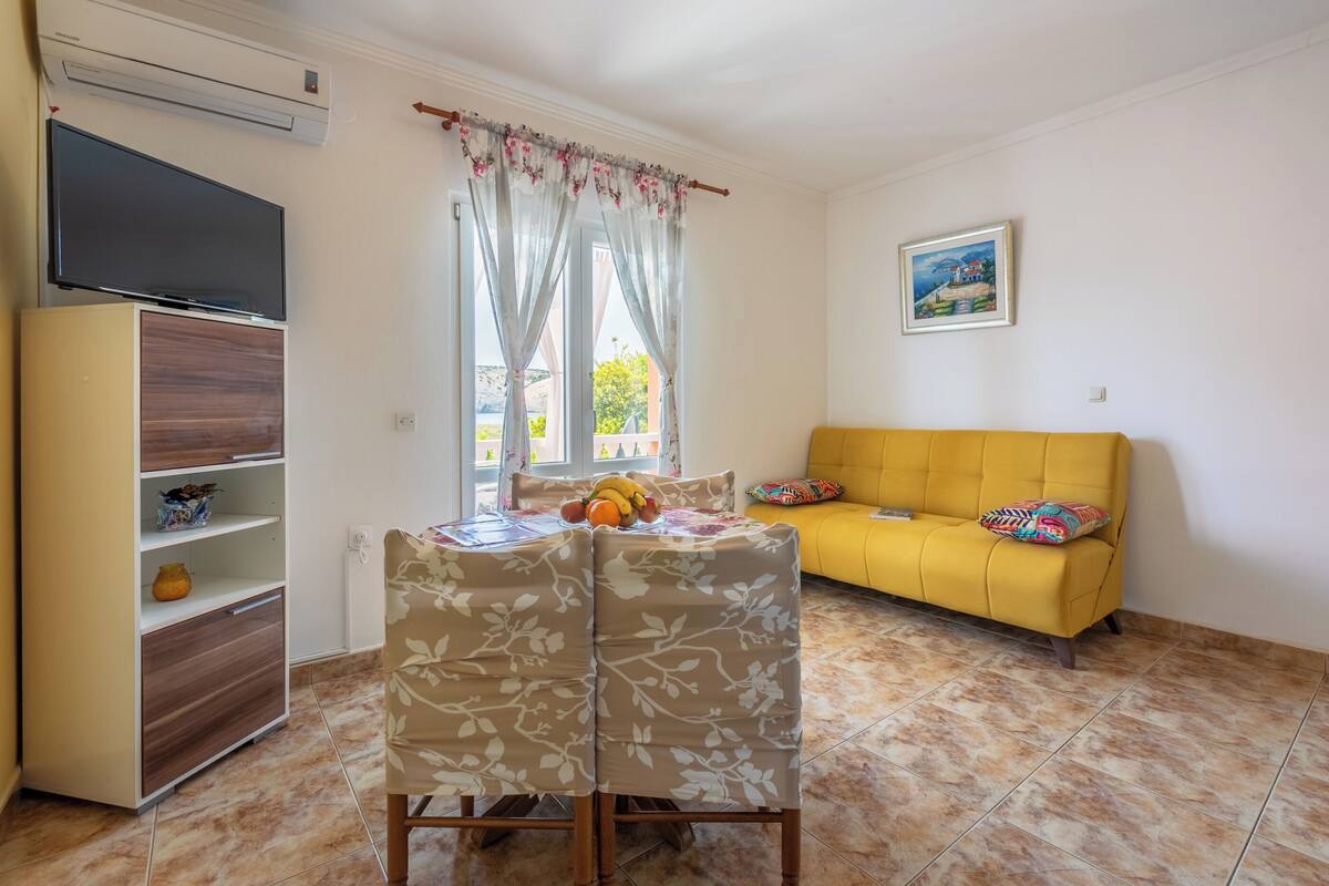 A room with a yellow sofa, a dining table with chairs, a TV on a cabinet, and a window with curtains.