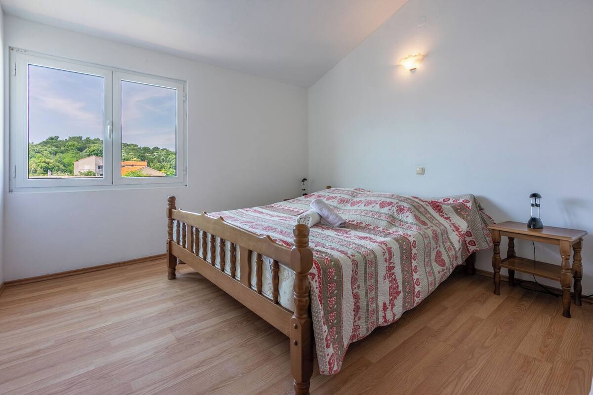 A bedroom with a wooden bed, a bedside table, and a window with a view of greenery.