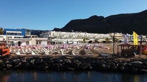 A beachside hotel with rows of sun loungers and umbrellas, a rocky shoreline, and buildings in the background. - Harbour 11 Apartment (Mogan)