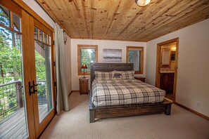A bedroom with a wooden ceiling, a bed with a plaid bedspread, a nightstand with a lamp, and a window with a view of greenery.