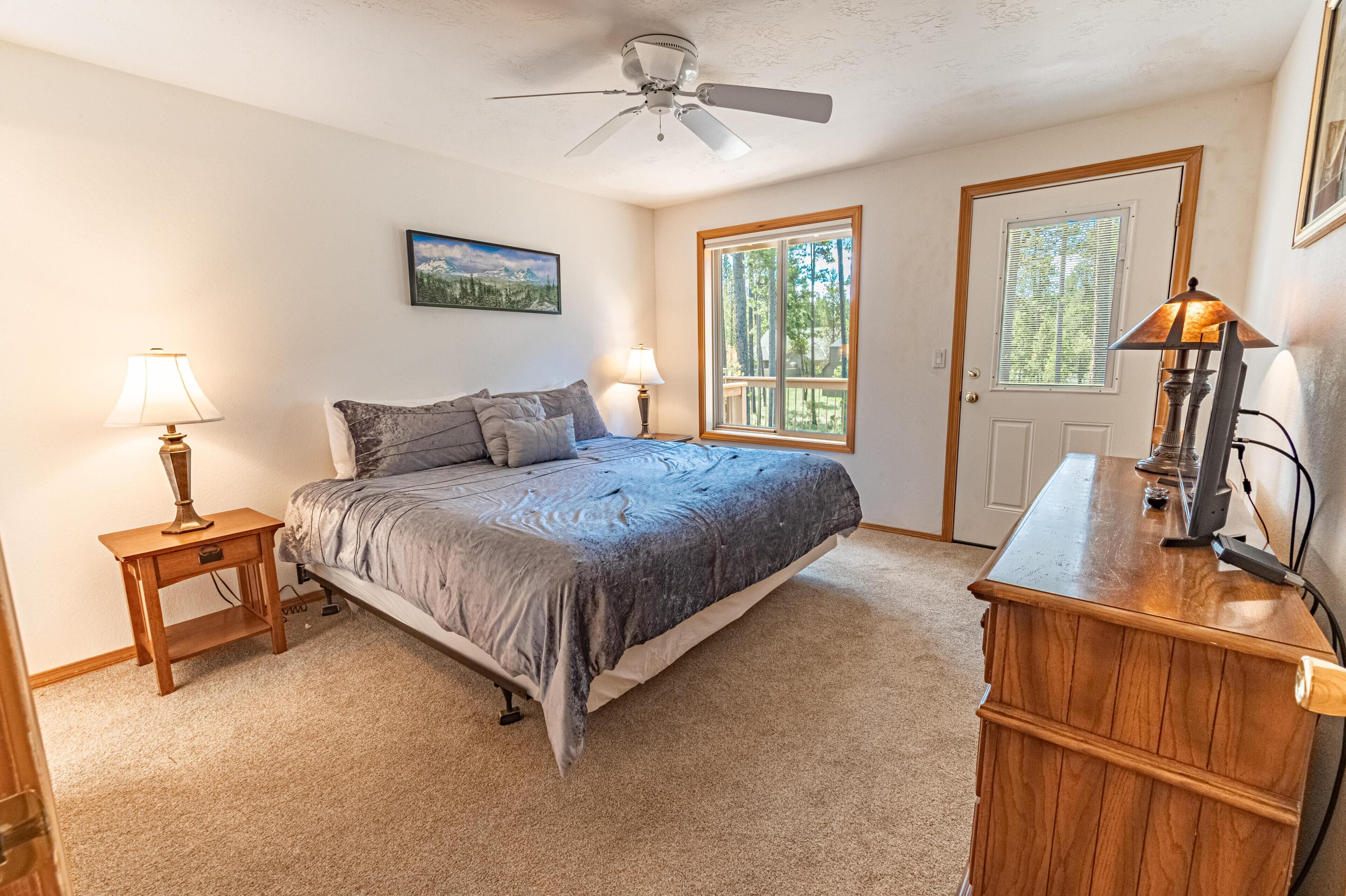 A bedroom with a bed, two bedside tables with lamps, a ceiling fan, a wooden dresser, and a door with a window.