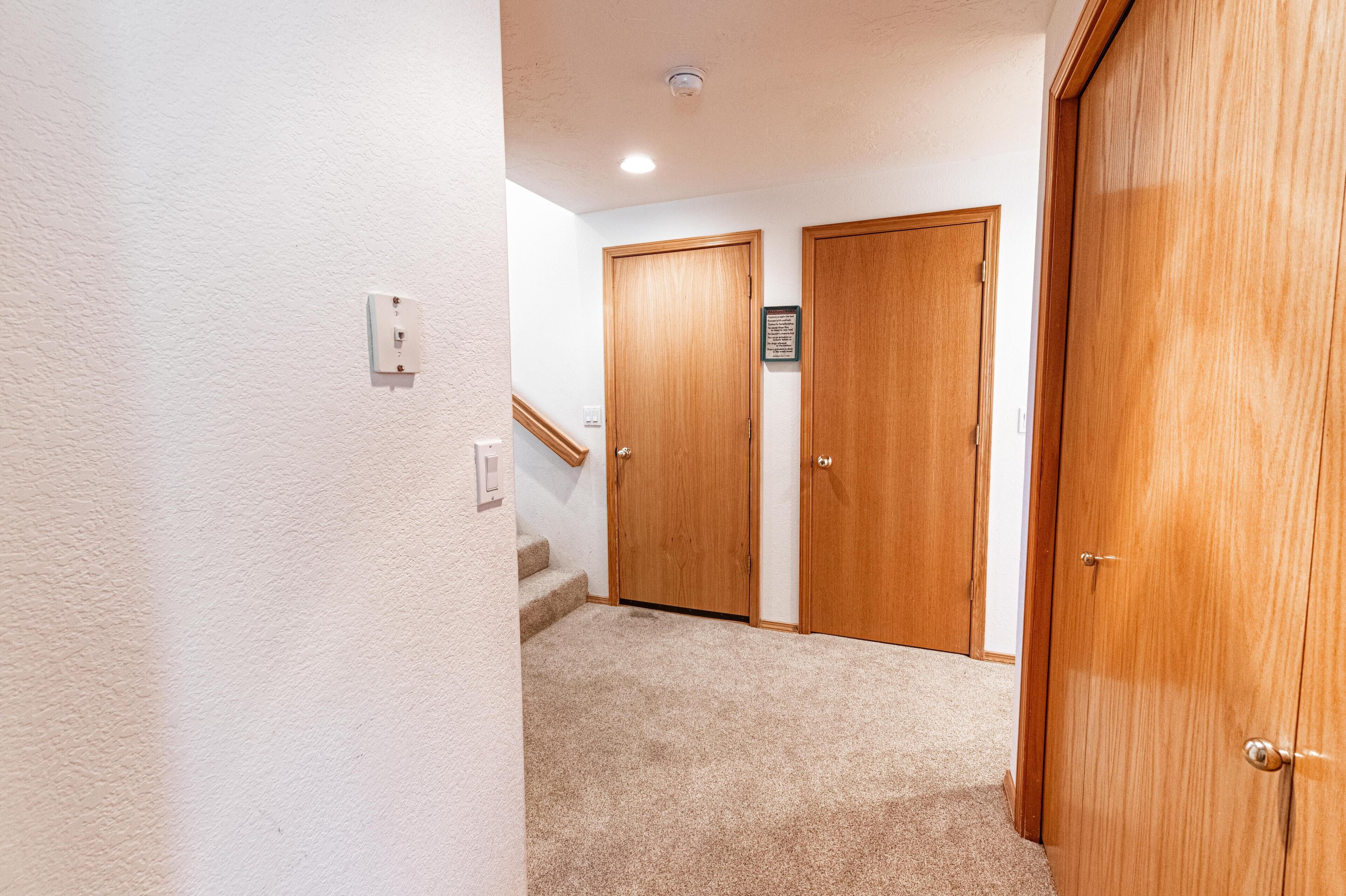 A hallway with carpeted flooring, wooden doors, a light switch, and a small sign on the wall.