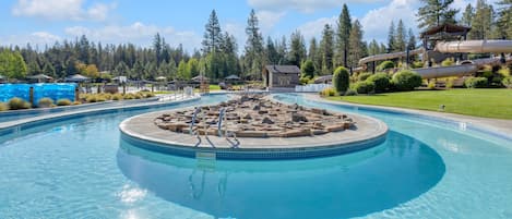 A large outdoor swimming pool with a central rock feature, surrounded by a deck and lounge areas, set in a forested area.