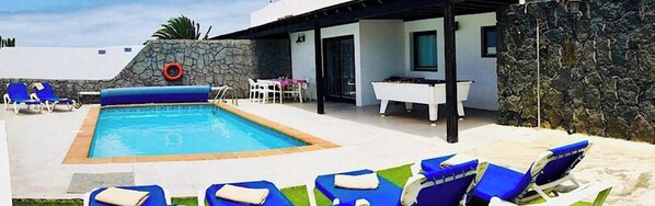 A modern outdoor pool area with blue and white lounge chairs, a stone wall, and a clear blue sky.