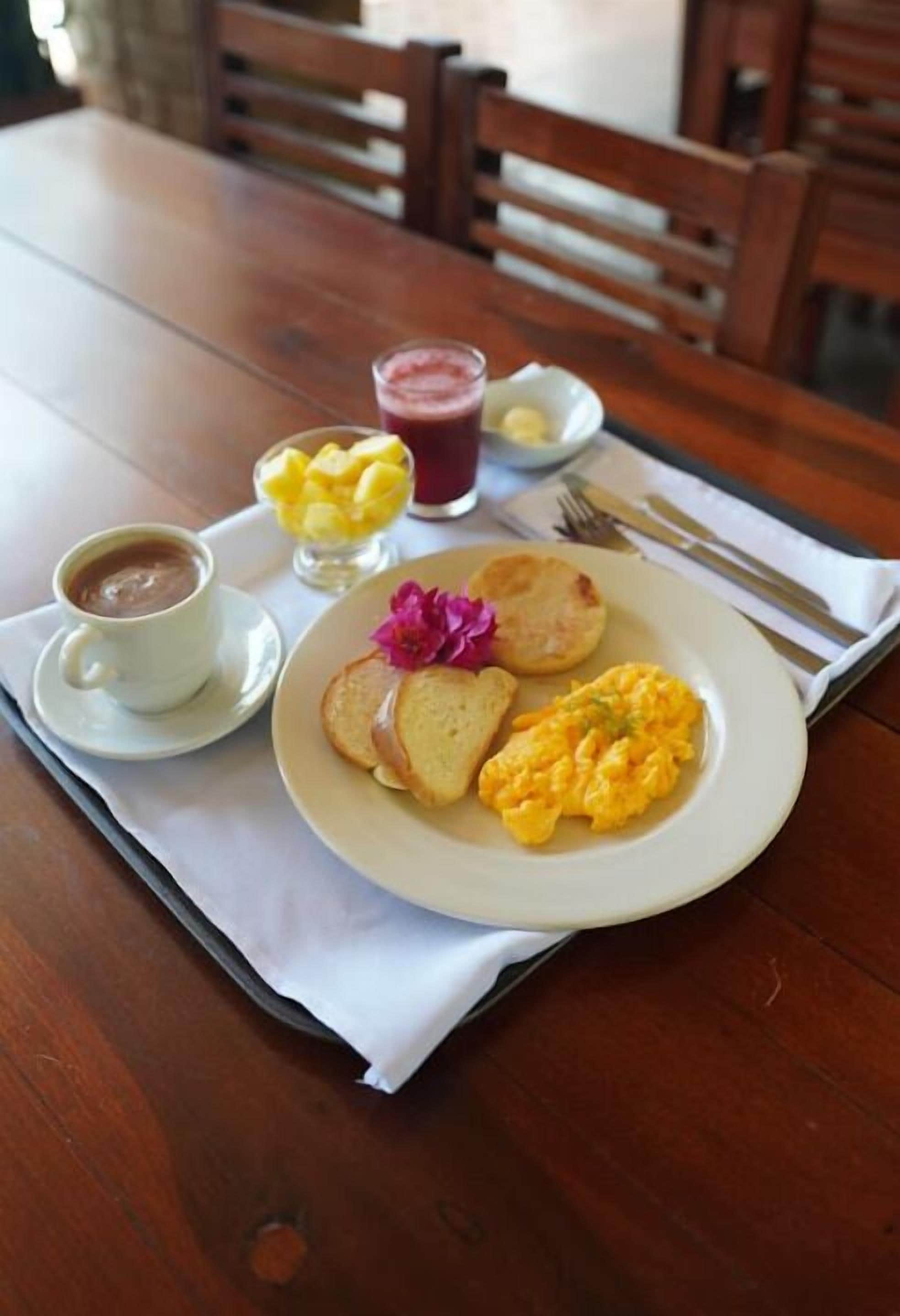 Uma bandeja de café da manhã com ovos mexidos, torradas, uma xícara de café e uma cesta de frutas sobre uma mesa de madeira.