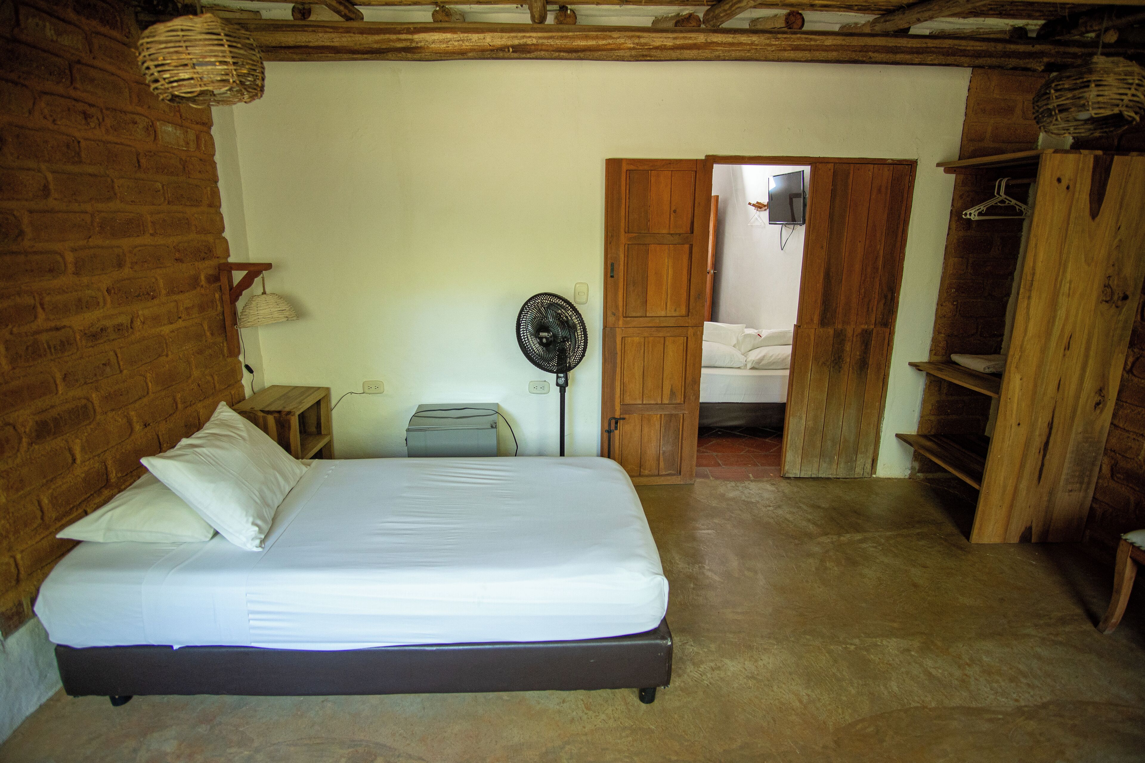 A bedroom with a bed, a fan, a wooden wardrobe, and a brick wall.