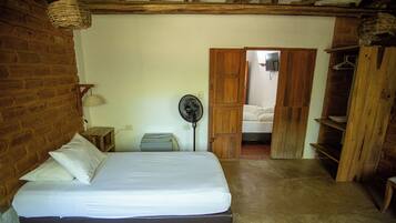 A bedroom with a bed, a fan, a wooden wardrobe, and a brick wall.