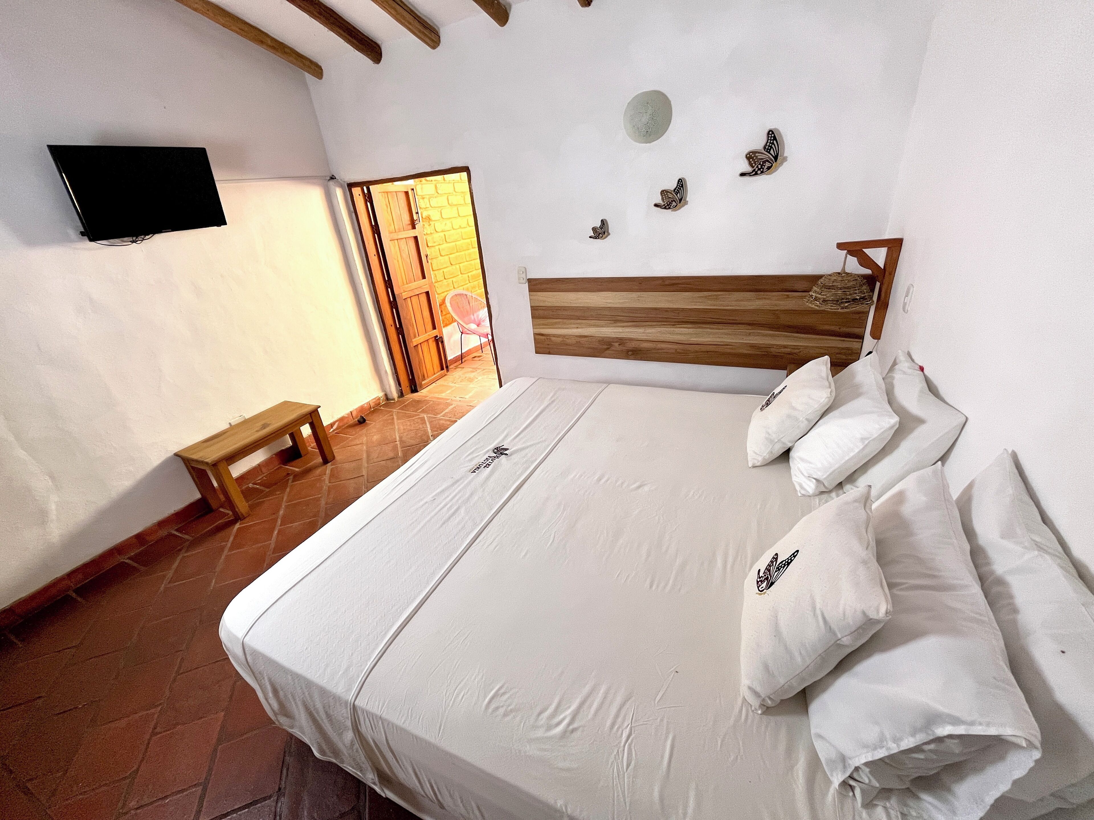 Un dormitorio con cama, almohadas, cabecera de madera, banco, televisor montado en la pared y puerta que da a un espacio exterior.