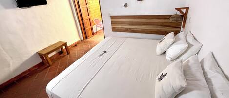 Un dormitorio con cama, almohadas, cabecera de madera, banco, televisor montado en la pared y puerta que da a un espacio exterior.