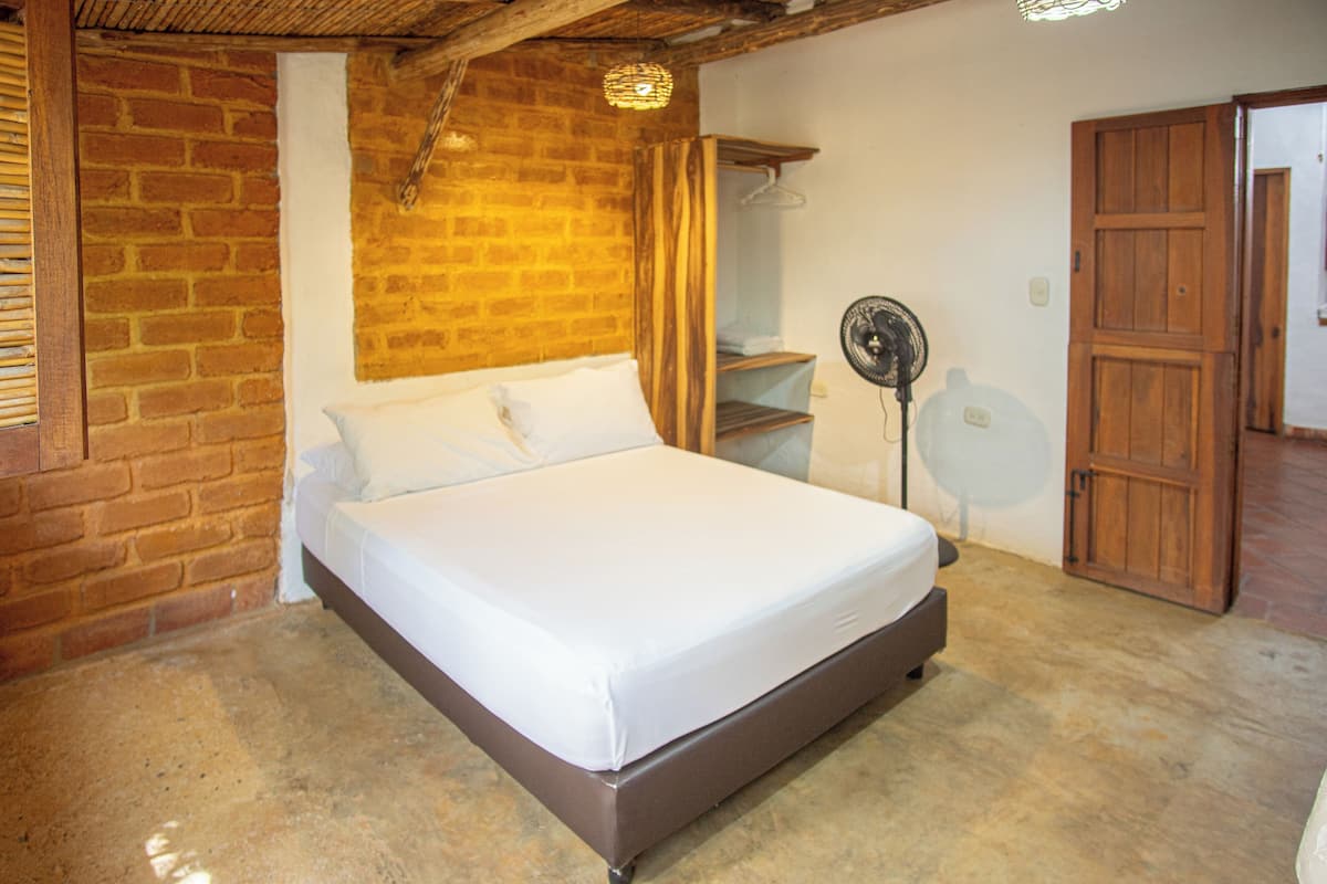 Ein Schlafzimmer mit einem großen Bett, Backsteinwänden, einer Holztür und einem Ventilator.