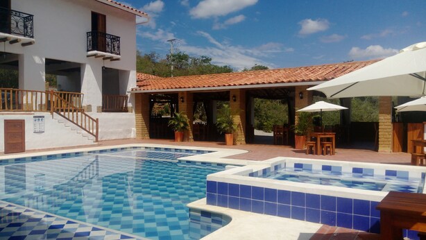 Ein Swimmingpool mit blauen und weißen Fliesen, eine überdachte Terrasse mit Sitzgelegenheiten im Freien sowie ein Gebäude mit einem gefliesten Dach und einem Balkon.