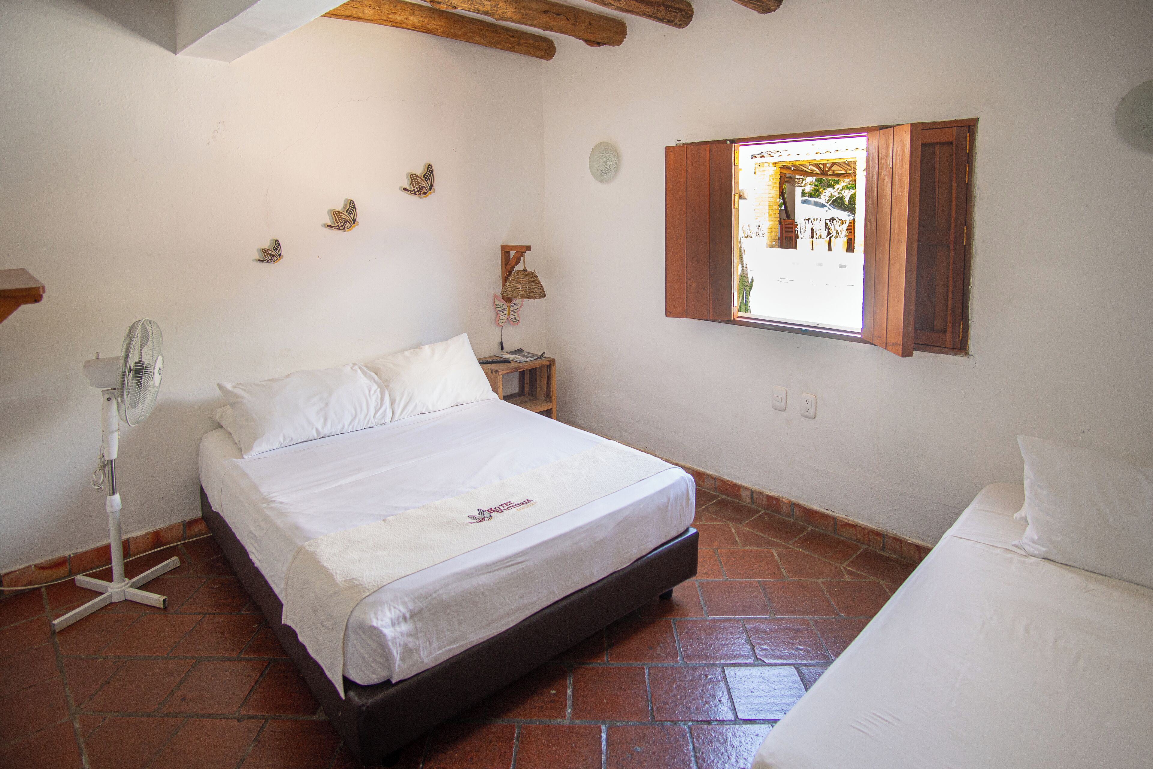 A bedroom with a bed, a bedside table, a lamp, a fan, and a window with wooden shutters.