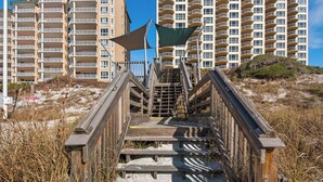 Una escalera de madera que lleva a una zona de playa con un letrero de salvavidas, rodeada de hierba alta y un edificio de gran altura al fondo.