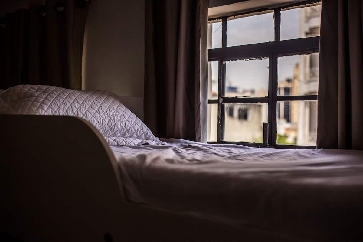 A bed with a quilted headboard, a window with a view of buildings, and curtains.