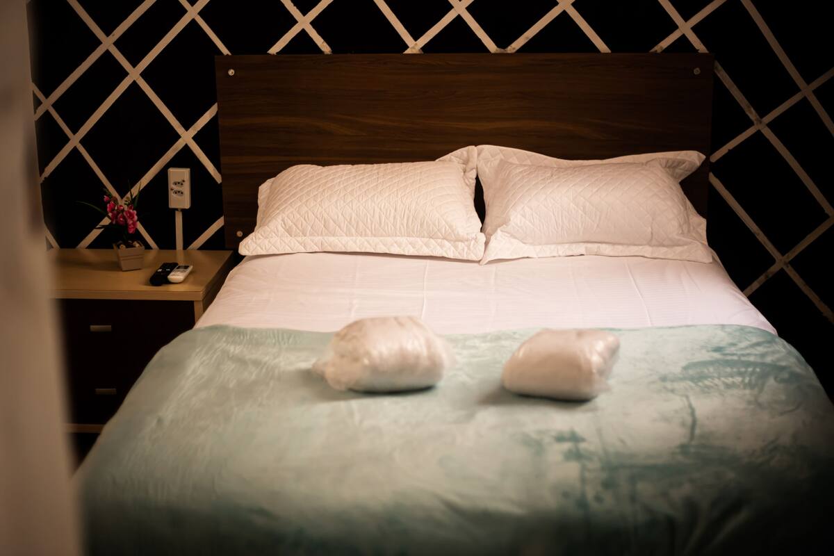 A neatly made bed with a quilted white duvet and pillows, a wooden headboard, and a nightstand with a vase of flowers and a remote control.