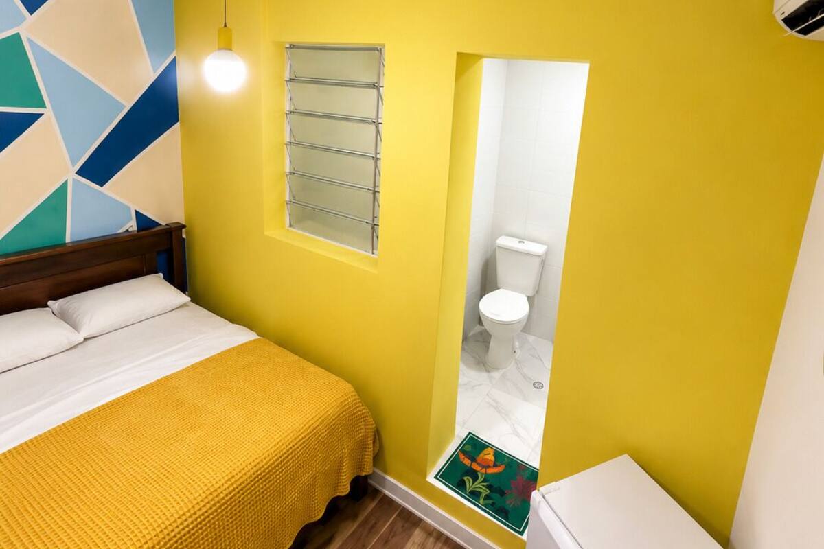 A small, brightly colored room with a bed, a compact refrigerator, and a bathroom visible through a doorway.