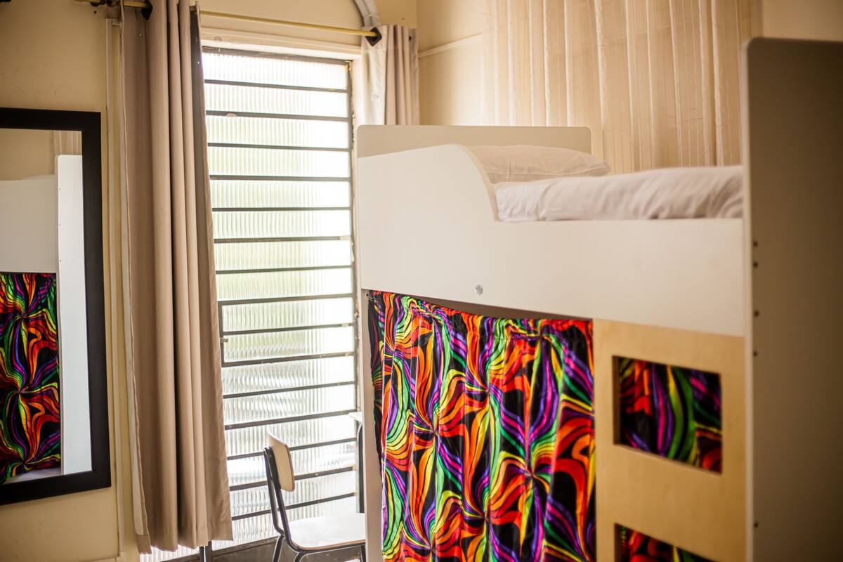 A room with a bunk bed, a chair, a window with blinds, and a colorful curtain.