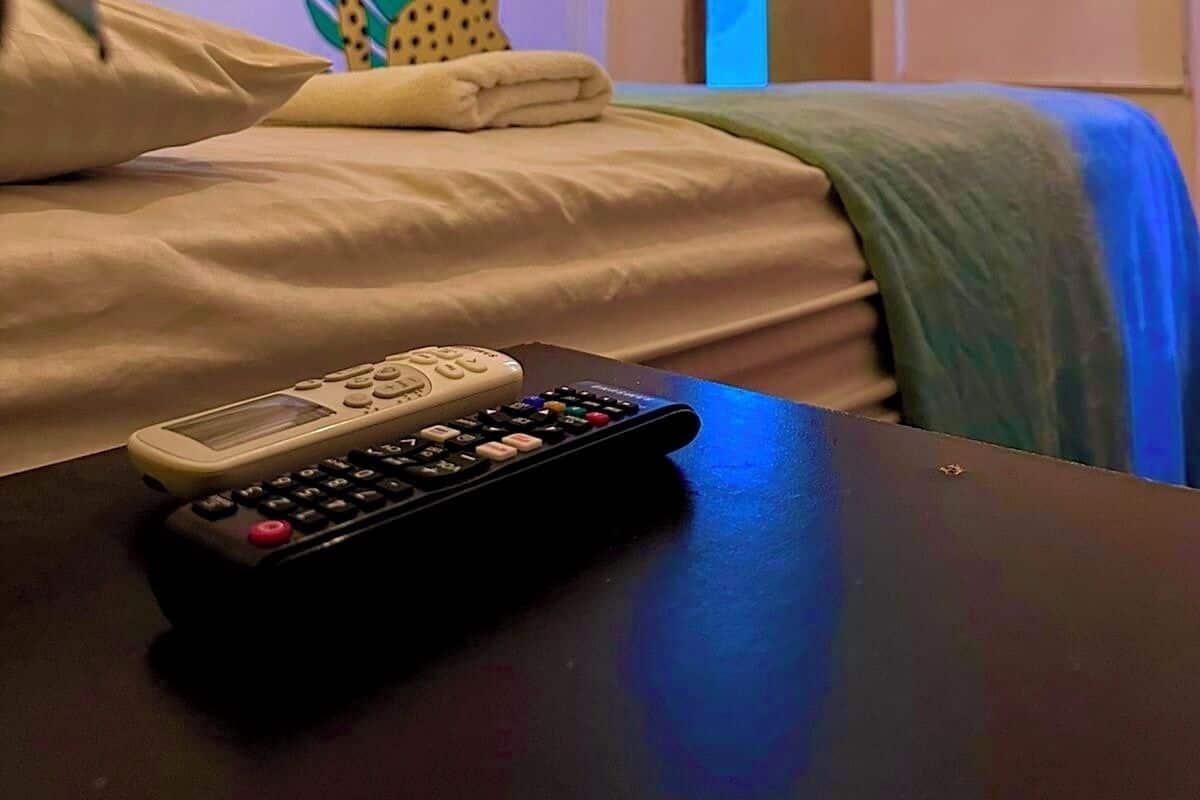 Habitación de hotel con cama, control remoto, aire acondicionado instalado en la pared y televisor mostrando una escena colorida.