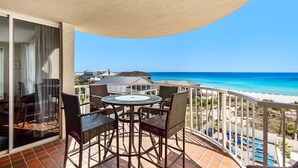 Ein Balkon mit Korbmöbeln und einer Glastür, mit Blick auf einen Strand mit klarem, blauem Wasser und einigen Gebäuden im Hintergrund.