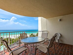 Un balcon avec une table ronde et des chaises, donnant sur une plage aux eaux bleu clair, où l’on aperçoit quelques personnes au loin.