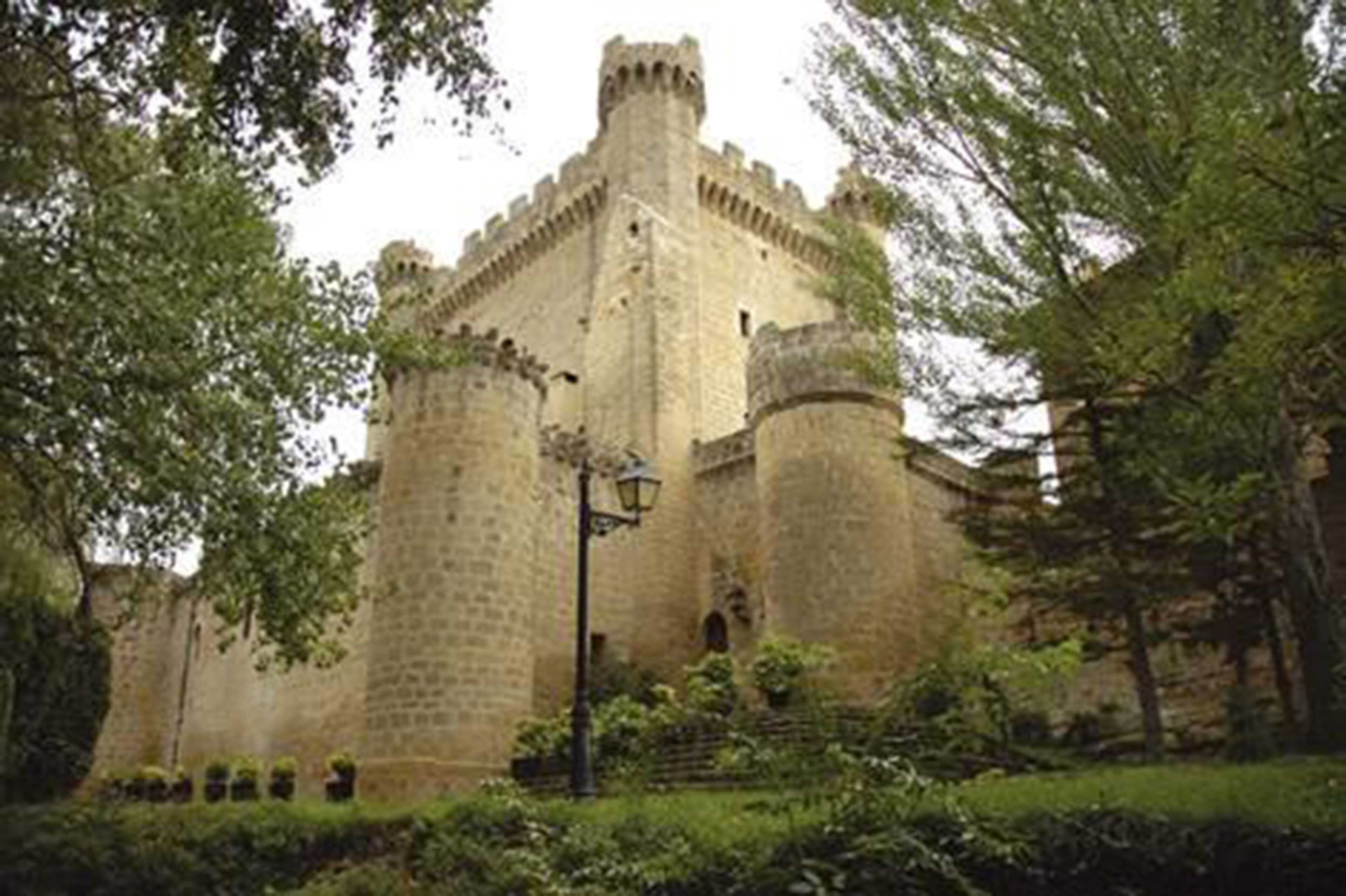 Un castell de pedra històric amb una torre central, envoltat de vegetació exuberante i un camí pavimentat.