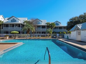 A swimming pool with a metal ladder, surrounded by a white fence, and adjacent to a building with balconies and palm trees.