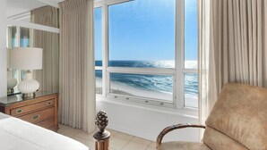 Ett hotellrum med ett stort fönster som vetter mot havet, en säng, en stol, en byrå och en lampa.