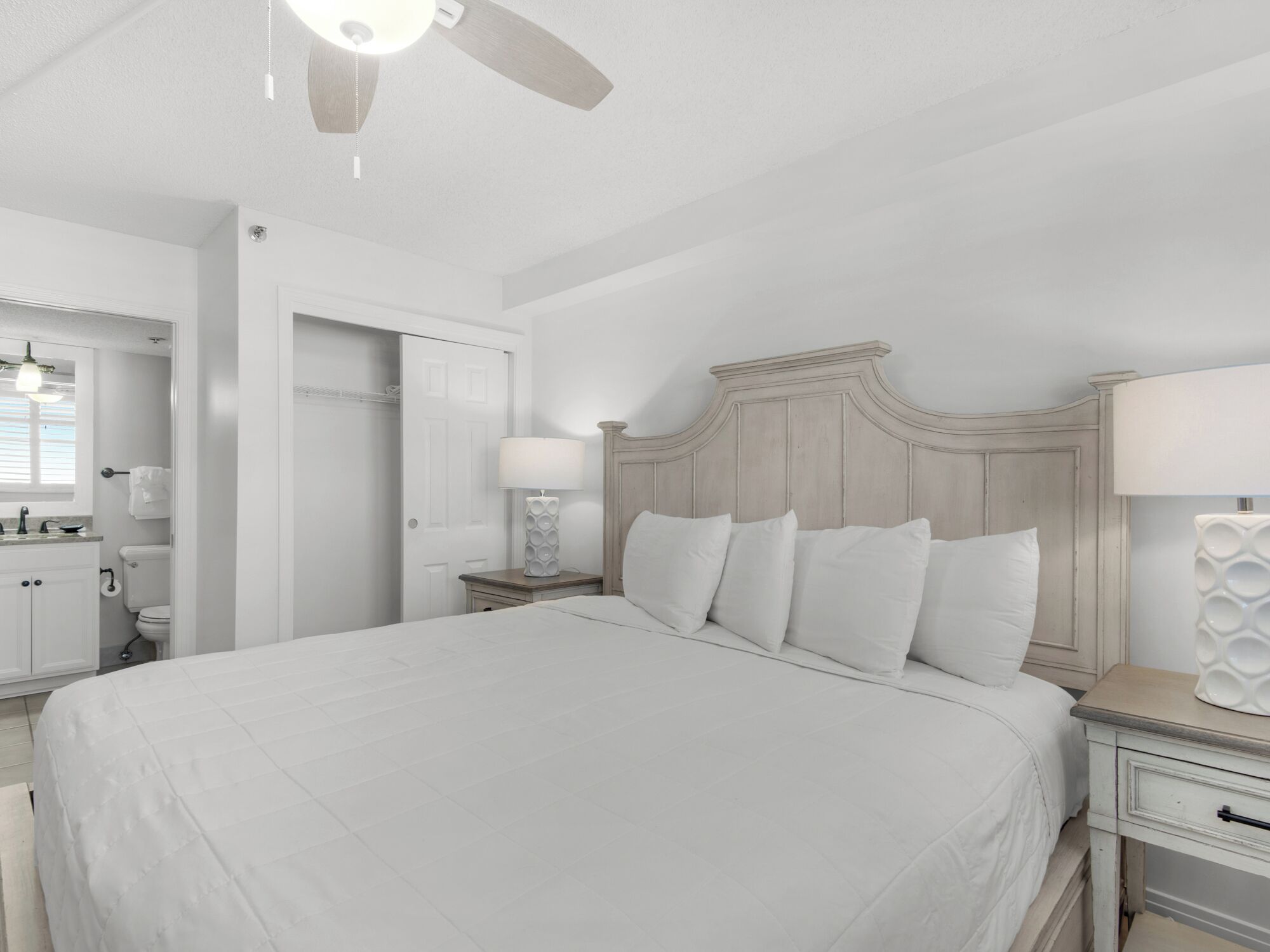 A bedroom with a large bed, a nightstand with a lamp, a ceiling fan, and a door leading to a bathroom.