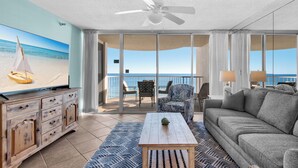 A spacious living room with a large TV, a ceiling fan, and a view of the ocean through large windows. - On the Beach, Pool, Huge TV, 3 Balconies, Umbrellas and Chairs Included (Miramar Beach)