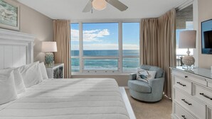 Une chambre d’hôtel avec un grand lit, un ventilateur de plafond, une vue sur l’océan, une chaise, une commode et une lampe.
