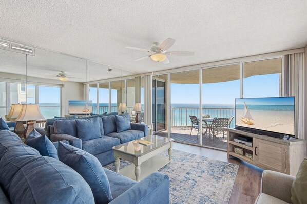 A spacious living room with a large sofa, a coffee table, and a television. There is a balcony with a view of the ocean.