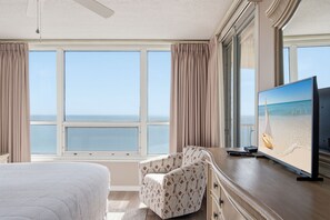 A hotel room with a large bed, a desk with a television, and a view of the sea through large windows.