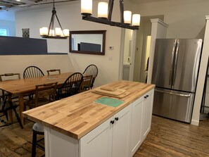 A kitchen with a wooden island, white cabinets, a stainless steel refrigerator, and a dining area with wooden chairs.