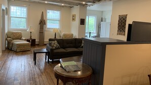 A living room with a wooden floor, a sofa, a chair, a small table, and a lamp. There are windows with blinds, a door, and a wall-mounted shelf. - Newly renovated in-town Bar Harbor, five bdrm apartment. Walk to shore & shops. (Bar Harbor)