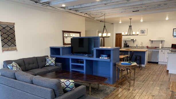 A modern living room with a grey sectional sofa, a small coffee table, and a TV on a blue entertainment unit. There is a kitchen area with a dining table and chairs, and a wooden floor throughout. - Newly renovated in-town Bar Harbor, five bdrm apartment. Walk to shore & shops. (Bar Harbor)