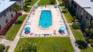 Vue aérienne d’un hôtel doté d’une piscine centrale entourée de chaises longues et de parasols, avec des bâtiments et une étendue d’eau en arrière-plan.