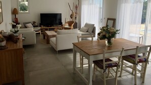 A modern living room with a flat-screen TV, sofas, a dining table with chairs, and a large window with sheer curtains. - New house with swimming pool and large outdoor terrace. (VENCE)