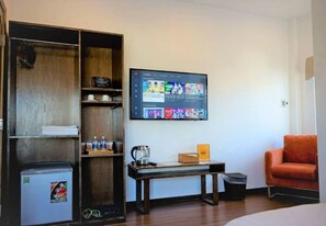 A hotel room with a wooden wardrobe, a small refrigerator, a TV mounted on the wall, a wooden table, and an orange chair.