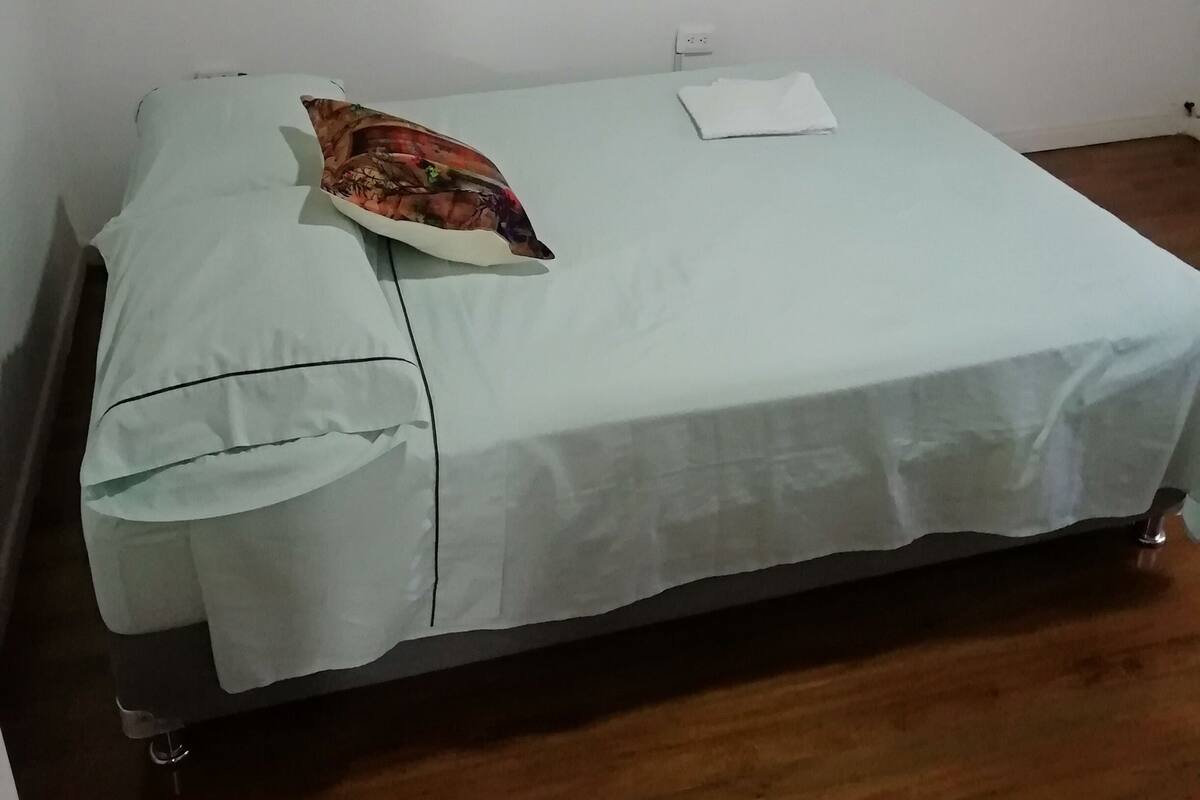 Un lit simple avec une couvre-lit de couleur claire et un coussin coloré, un petit tableau encadré accroché au mur et une prise de courant.