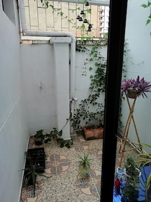 Un pequeño balcón cerrado con plantas en macetas, piso de baldosas y vista a un edificio al fondo.