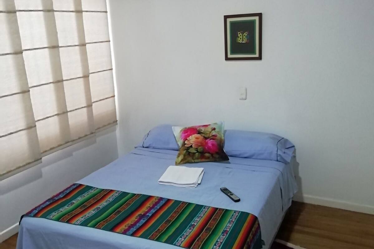 Une chambre à coucher avec un lit, une couverture colorée, une télécommande et un tableau encadré accroché au mur.