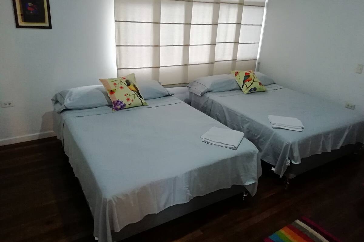 Une chambre avec deux lits simples, chacun muni d’un oreiller et d’une serviette pliée. Il y a une fenêtre avec des stores, un tableau encadré au mur et un plancher en bois.