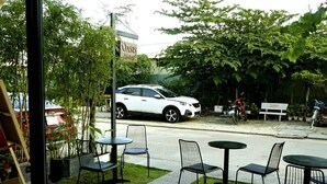 Ein weißer SUV parkt vor einem Gebäude, an dem ein Schild mit der Aufschrift "OASIS Hotel" angebracht ist. Im Freien stehen Tische und Stühle bereit, und ein Motorrad befindet sich in der Nähe.