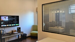Ein modernes Wohnzimmer mit einem an der Wand befestigten Flachbildfernseher, einem bequemen grünen Sessel, einem kleinen Holztisch mit Korb und einem großen Fenster mit Jalousien.