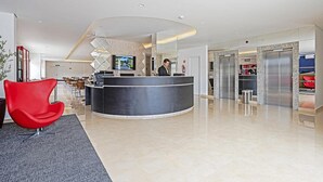Een moderne hotellobby met een receptiebalie, een rode stoel en een televisiescherm.