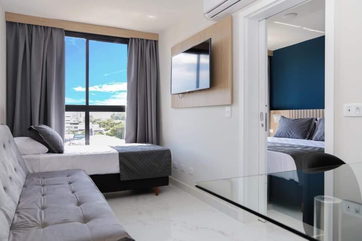 Une chambre d’hôtel moderne dotée d’une grande fenêtre, d’un téléviseur à écran plat fixé au mur, d’un canapé et d’un lit avec une tête de lit.