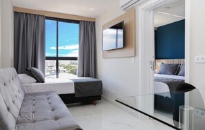 A modern hotel room with a large window, a flat-screen TV mounted on the wall, a sofa, and a bed with a headboard. - Slim João Bettega (Curitiba)