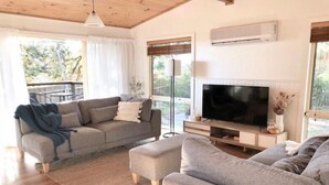 A living room with a grey sofa, a television, and a large window. - Bush Hideaway (Binalong Bay)