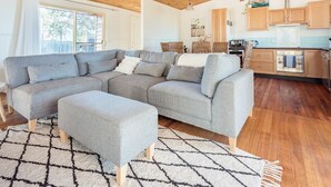 A modern living room with a grey sectional sofa, a matching ottoman, and a geometric rug. A kitchen with wooden cabinets and stainless steel appliances is visible in the background. - Bush Hideaway (Binalong Bay)