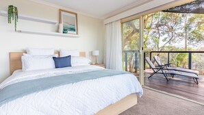 A bedroom with a large bed, a balcony with a view of trees, and a small table with a lamp. - Bush Hideaway (Binalong Bay)